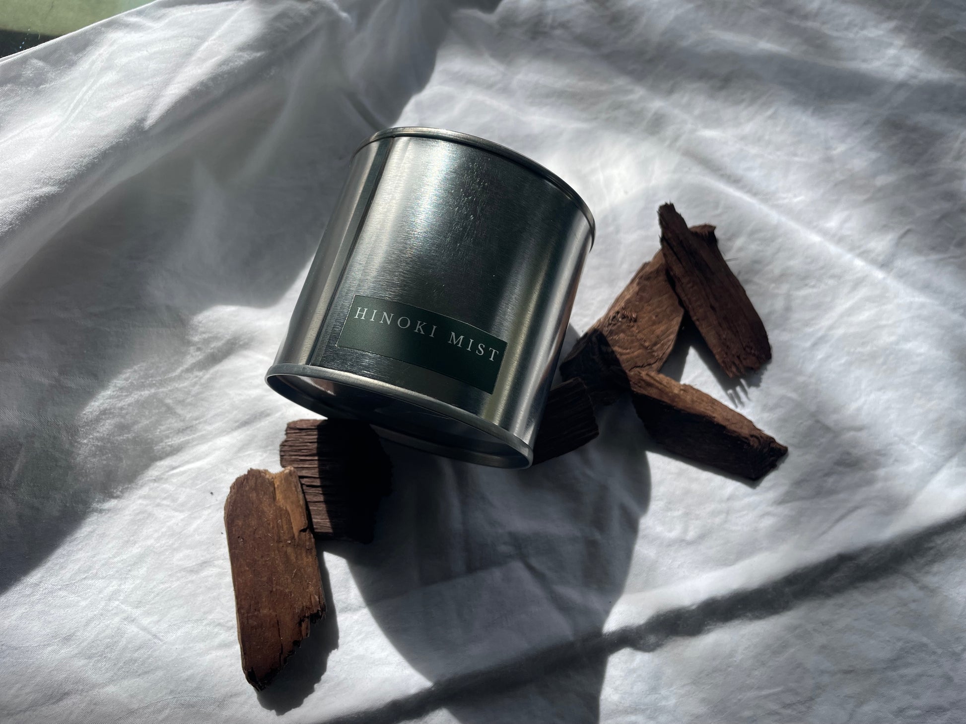 Hinoki Mist 03 candle by HOT WAX in steel tin on white fabric with hinoki wood pieces